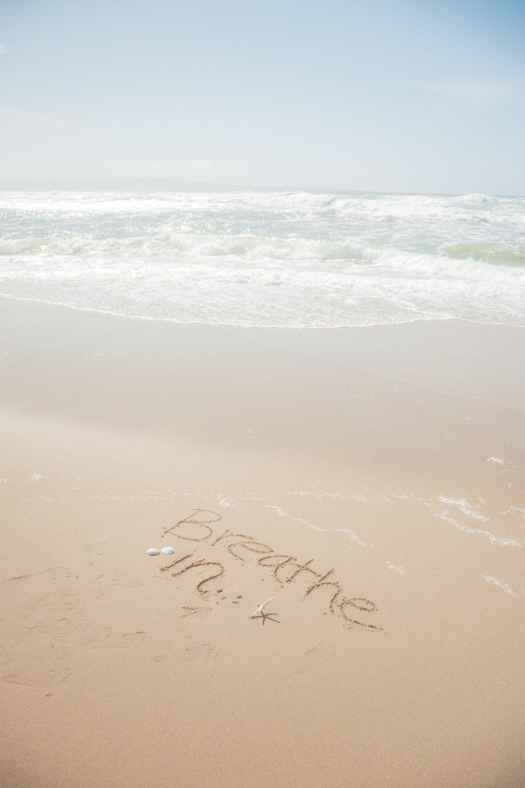 Breath in written in the sand with a sea in the background in open blue sky