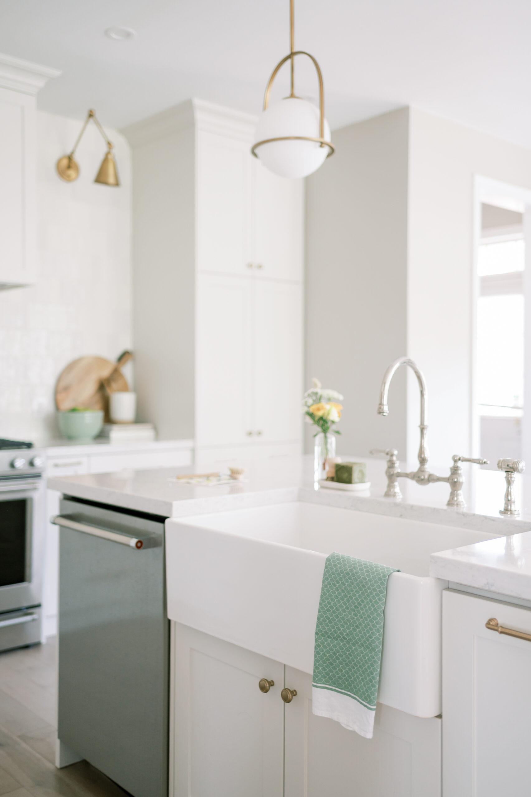 A bright, white modern kitchen featuring a farmhouse sink with a green towel, white cabinets, and a hanging pendant light. stainless steel appliances and marble countertops are visible.