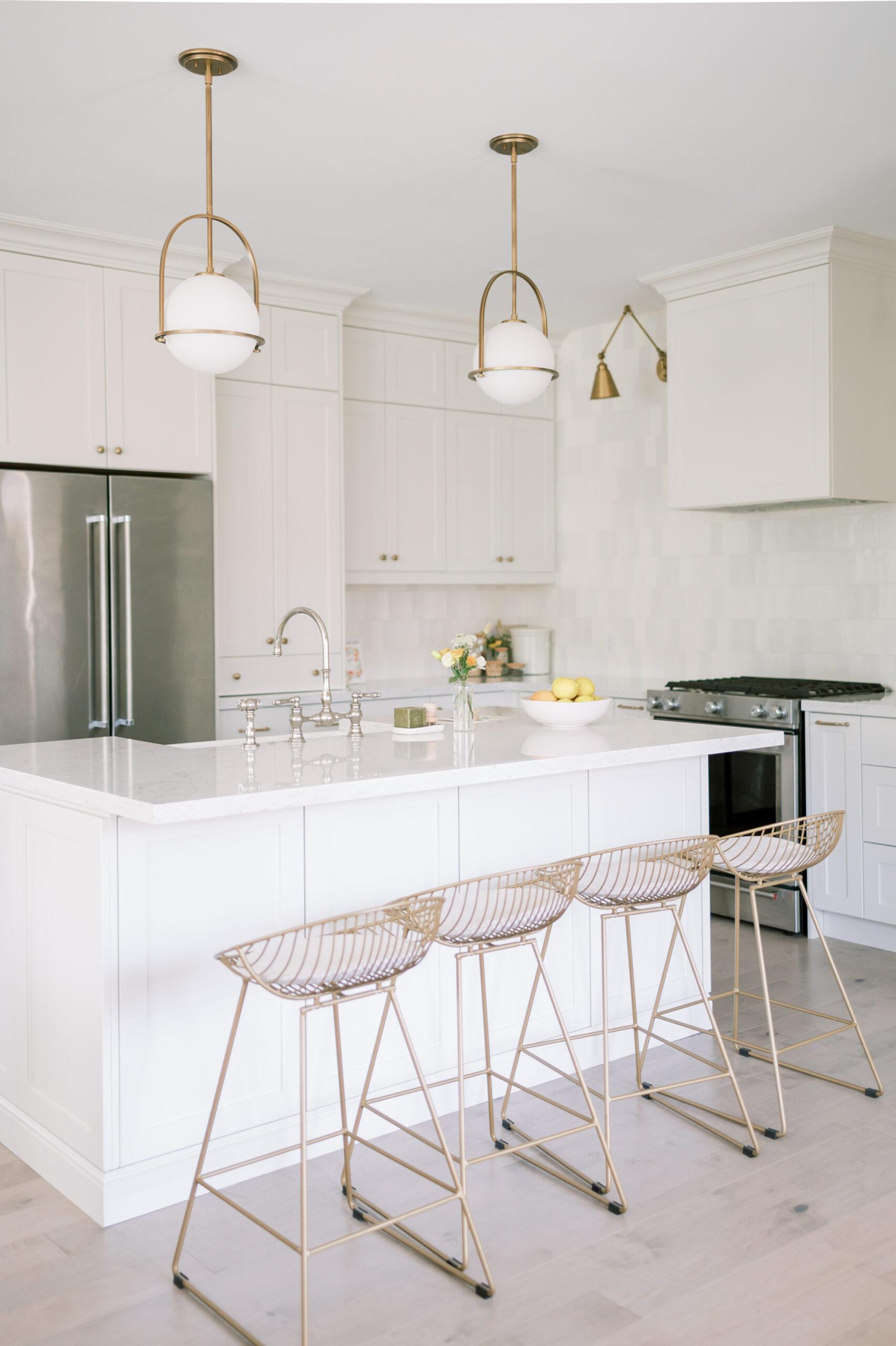 A bright, modern kitchen with white cabinetry, marble countertops, and gold accented bar stools at the island. pendant lights hang above, and a bouquet of flowers adds a touch of color.