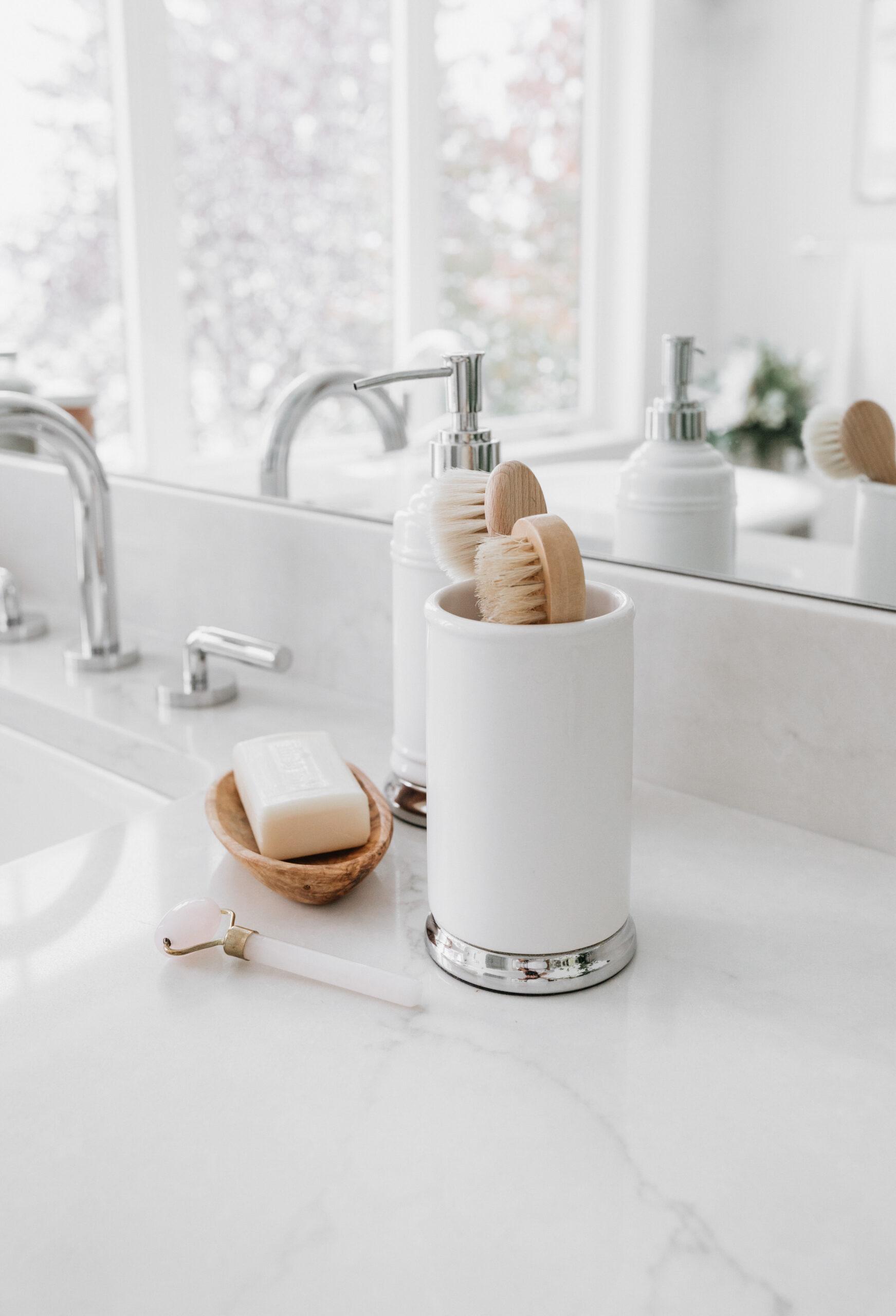 Elegant bathroom countertop with a marble surface, featuring a silver faucet, a white and silver cylindrical container holding wooden brushes, soap, and a facial roller. bright, natural light fills the space.