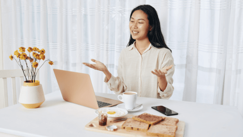 A girl sitting on a table with a laptop and a tray of breakfast  and tea is form of her and there is also a vase  with white curtains in the background