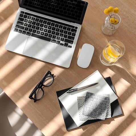 Laptop, a wireless mouse, a vase with yellow flowers,  in it , a glass of lie infused water, a pair of glasses and  pen and paper